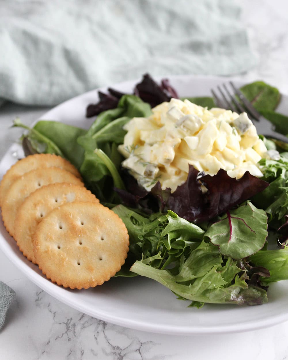 a scoop of egg salad on a bed of field greens on a white plate with crackers