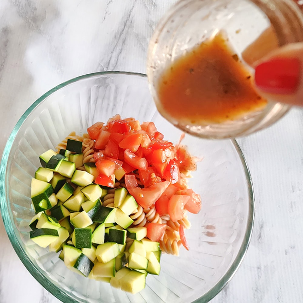 a picture of the pasta salad in a bowl with dressing pouring in